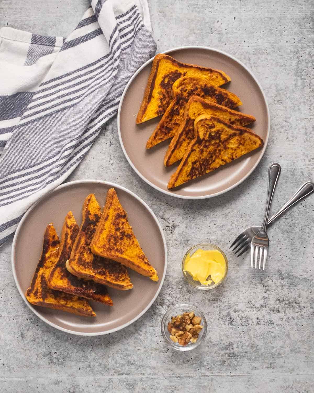 Two plates with pumpkin French toast wedges, two forks, butter, and chopped walnuts.
