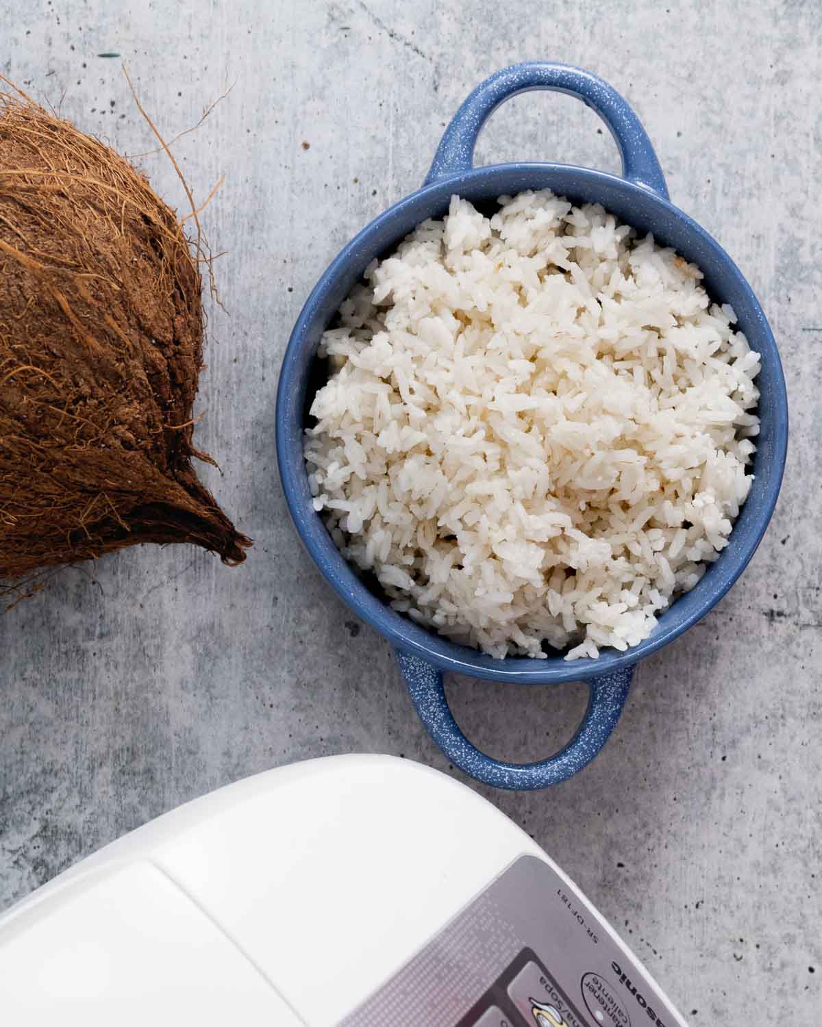 A top view of a bowl with cooked coconut rice in a bowl, a coconut and a rice cooker on the side.