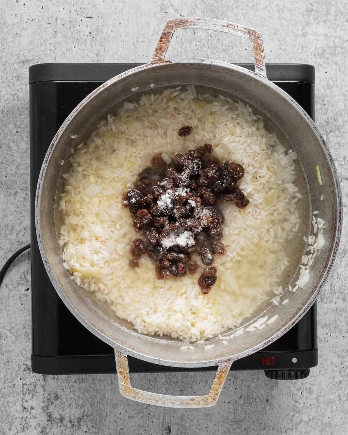 Before cooking, a pot with sautéed onion, garlic, rice, raisins, salt, sugar, and water.
