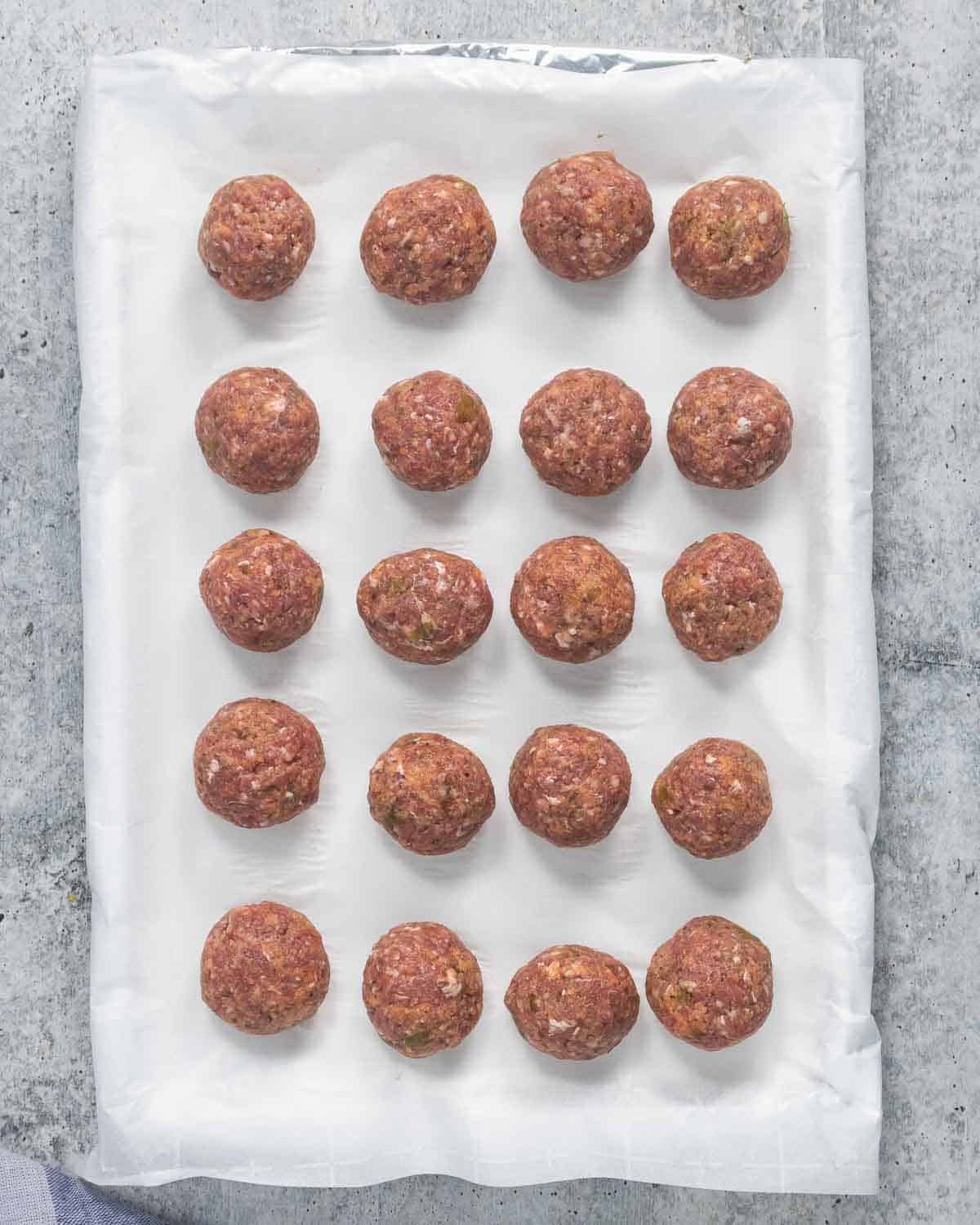 Twenty raw beef meatballs on a lined parchment paper baking sheet.